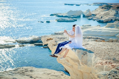 女人穿着婚纱坐在男人穿着白色礼服衬衫在石头尖面临大海
